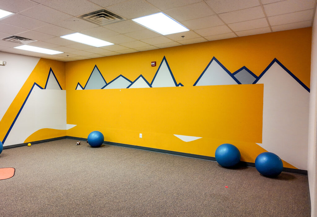 Carpeted flexible-use room with open floor area and geometric wall mural
