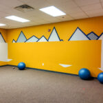 Carpeted flexible-use room with open floor area and geometric wall mural